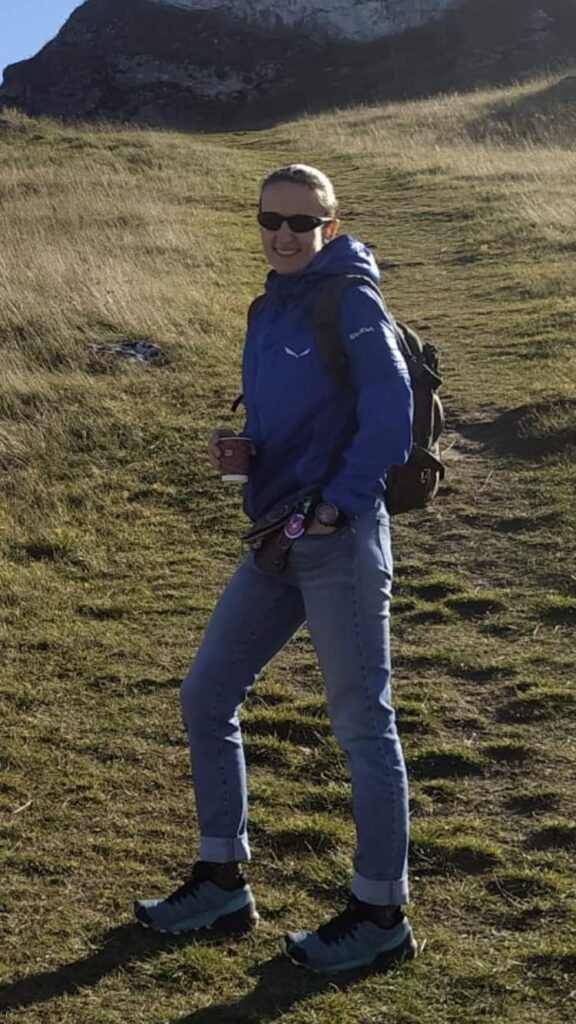 Płk rez. dr Izabela Wlizło w niebieskiej kurtce sportowej i z plecakiem podczas górskiej wędrówki. Ekspertka od budowania zespołów i autorka książki stoi uśmiechnięta na tle górskiego szlaku, trzymając kubek z kawą