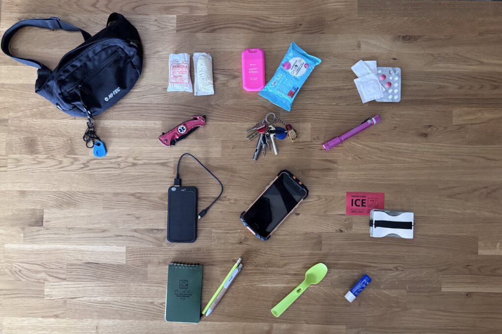 Zdjęcie typu flat lay przedstawiające zestaw Everyday Carry (EDC) ułożony na drewnianym blacie. Widoczna czarna nerka Hi-Tec oraz uporządkowane przedmioty: medyczne (maseczka RKO, rękawiczki, leki), techniczne (telefon, powerbank, latarka), scyzoryk ratowniczy, klucze z gwizdkiem oraz notes z długopisem i zielona łyżeczka