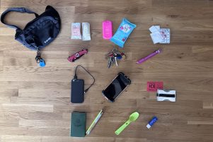 Zdjęcie typu flat lay przedstawiające zestaw Everyday Carry (EDC) ułożony na drewnianym blacie. Widoczna czarna nerka Hi-Tec oraz uporządkowane przedmioty: medyczne (maseczka RKO, rękawiczki, leki), techniczne (telefon, powerbank, latarka), scyzoryk ratowniczy, klucze z gwizdkiem oraz notes z długopisem i zielona łyżeczka